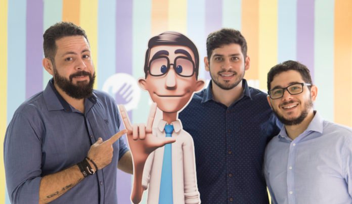 Descrição da imagem #PraCegoVer: Imagem no formato retangular, na horizontal. Os fundadores da Hand Talk com o Hugo. À esquerda está Carlos, um homem branco de cabelos curtos e barba, ele veste camisa azul. Ao seu lado, o Hugo, fazendo o sinal de 