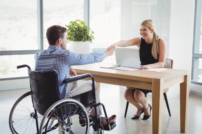 Descrição da imagem #PraCegoVer: Imagem no formato retangular, na horizontal. Um rapaz cadeirante está cumprimentando uma mulher. Entre eles tem uma mesa, de madeira, com um notebook em cima. Fim da descrição.