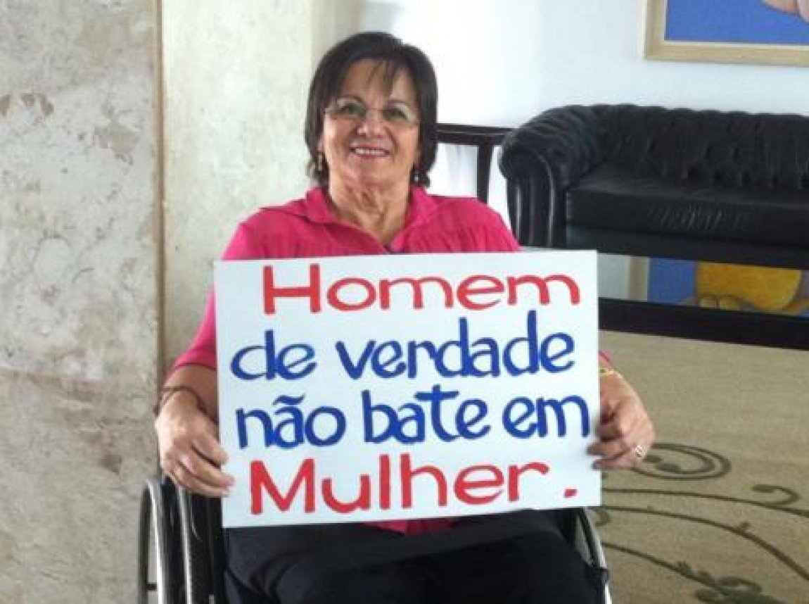 Descrição de imagem #PraCegoVer: Imagem no formato retangular, na horizontal. Maria da Penha está sentada em sua cadeira de rodas. Ela segura um cartaz onde está escrito: Homem de verdade não bate em mulher. Fim da descrição.