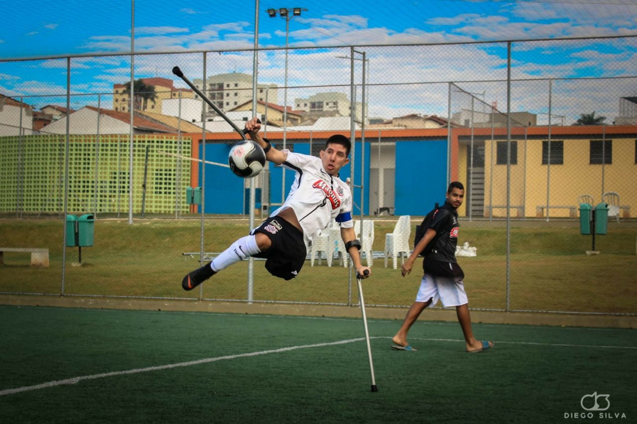 Descrição da imagem #PraCegoVer: Imagem no formato retangular, na horizontal. O jogador Rogerinho, durante lance de um jogo. Ele está apoiado em uma de suas muletas e tem o corpo levantado, para chutar a bola. Fim da descrição.