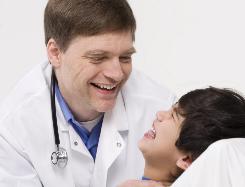 Descrição da imagem #PraCegoVer: Imagem no formato retangular na horizontal. Um médico está sorrindo, ao lado de um menino, deitado em uma cama de hospital. O menino também está sorrindo. Fim da descrição.