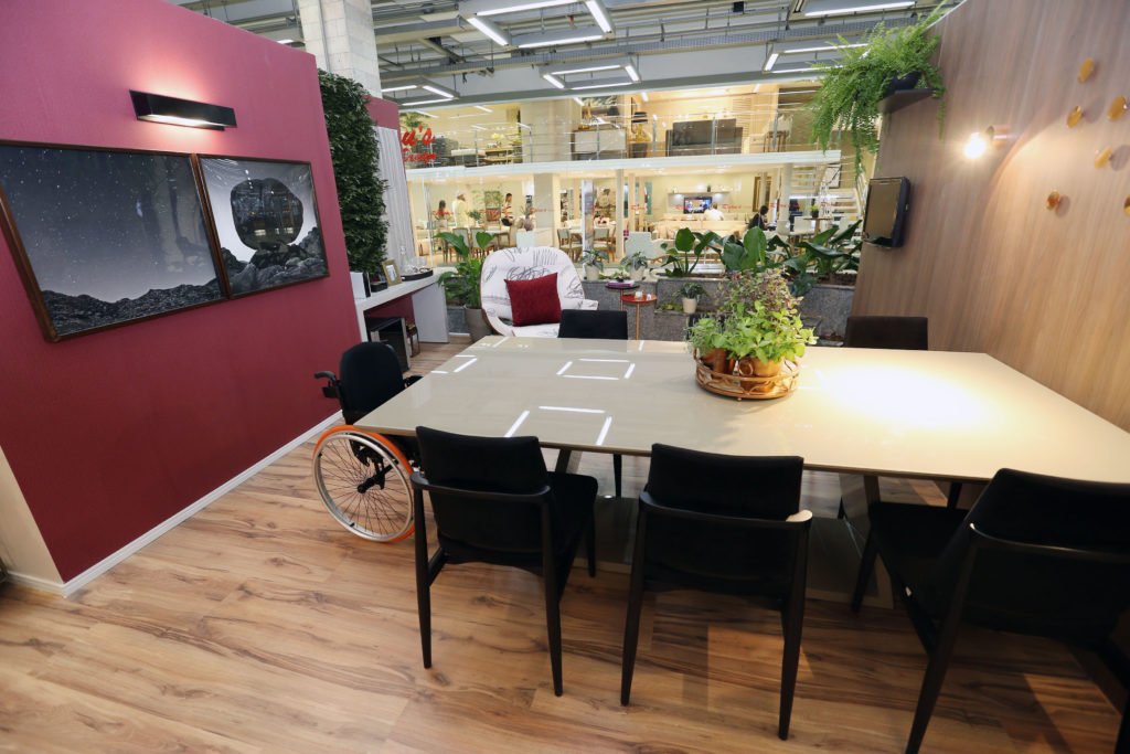 Descrição da imagem #PraCegoVer: Imagem no formato retangular, na horizontal. Sala de jantar e varanda gourmet, da mostra de arquitetura e desenho universal do shopping Lar Center. No ambiente vemos, em primeiro plano, uma mesa branca com um arranjo de folhas ao centro, e seis cadeiras pretas. Na ponta da mesa, temos uma cadeira de rodas. Uma das paredes é revestida em madeira e outra é evidenciada pela cor marsala. Nela estão dois quadros, com molduras pretas. Ao fundo vemos uma poltrona branca, com uma almofada vermelha, e uma TV na parede. Fim da descrição.