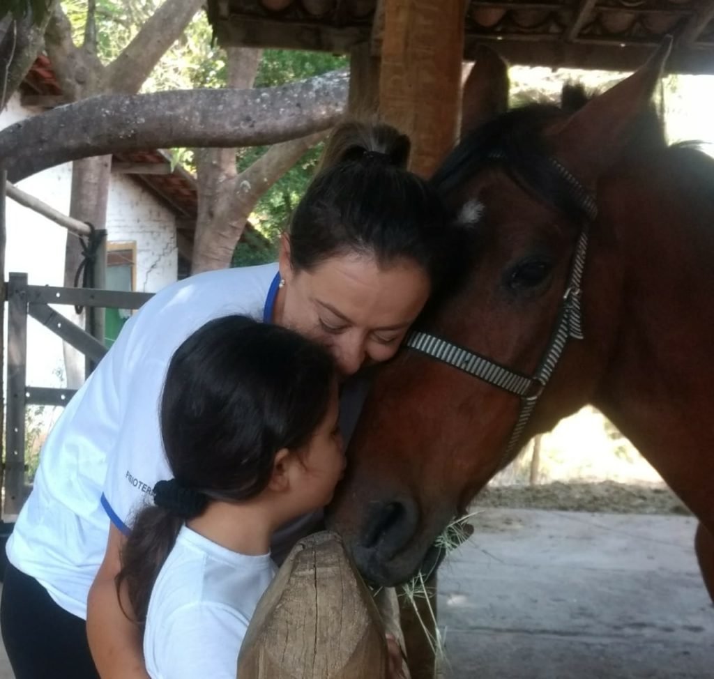 Descrição da imagem #PraCegoVer: Imagem no formato quadrado. Uma menina com autismo está interagindo com o cavalo, no setting de equoterapia. A menina está acompanhada de uma profissional. Fim da descrição.