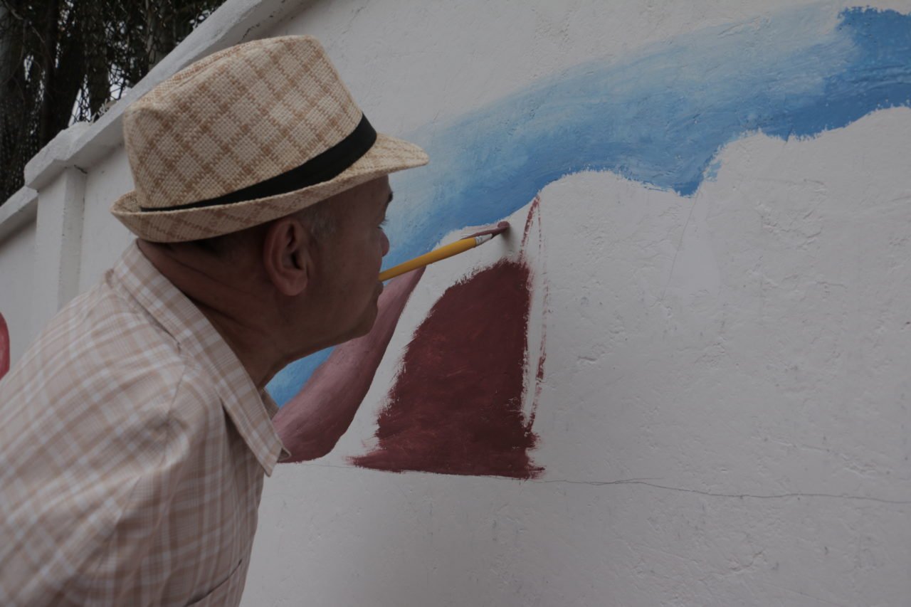 Descrição da imagem #PraCegoVer: A imagem está no formato retangular, na horizontal. Um homem com deficiência está segurando um pincel com a boca e pinta um muro branco. Fim da descrição.