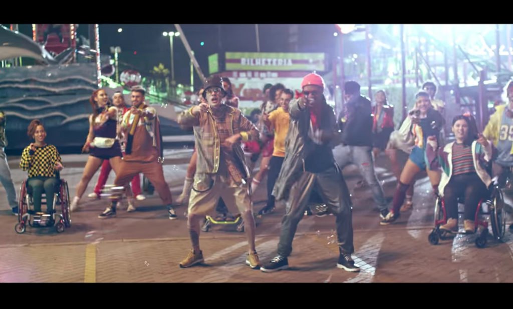 Descrição da imagem #PraCegoVer: Imagem no formato retangular, na horizontal. Cena do clipe de O Bebê, com Kevinho e MC Kekel. Eles estão em um parque de diversões, com várias luzes coloridas, ao fundo. Atrás deles, um grupo de pessoas estão dançando, uma coreografia. Tem pessoas cadeirantes. Fim da descrição.