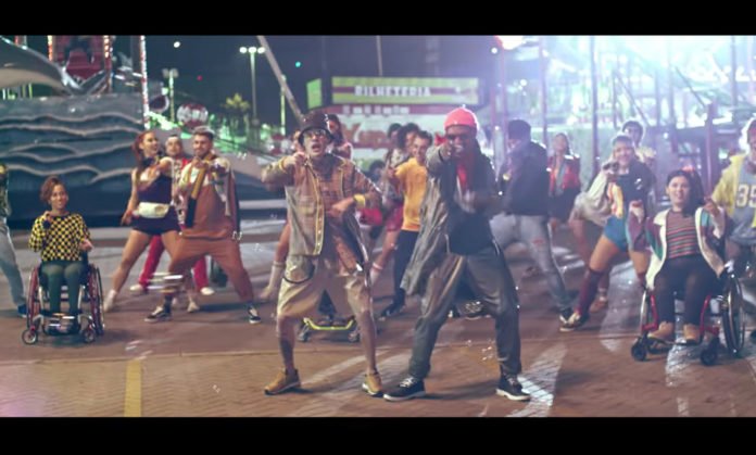 Descrição da imagem #PraCegoVer: Imagem no formato retangular, na horizontal. Cena do clipe de O Bebê, com Kevinho e MC Kekel. Eles estão em um parque de diversões, com várias luzes coloridas, ao fundo. Atrás deles, um grupo de pessoas estão dançando, uma coreografia. Tem pessoas cadeirantes. Fim da descrição.