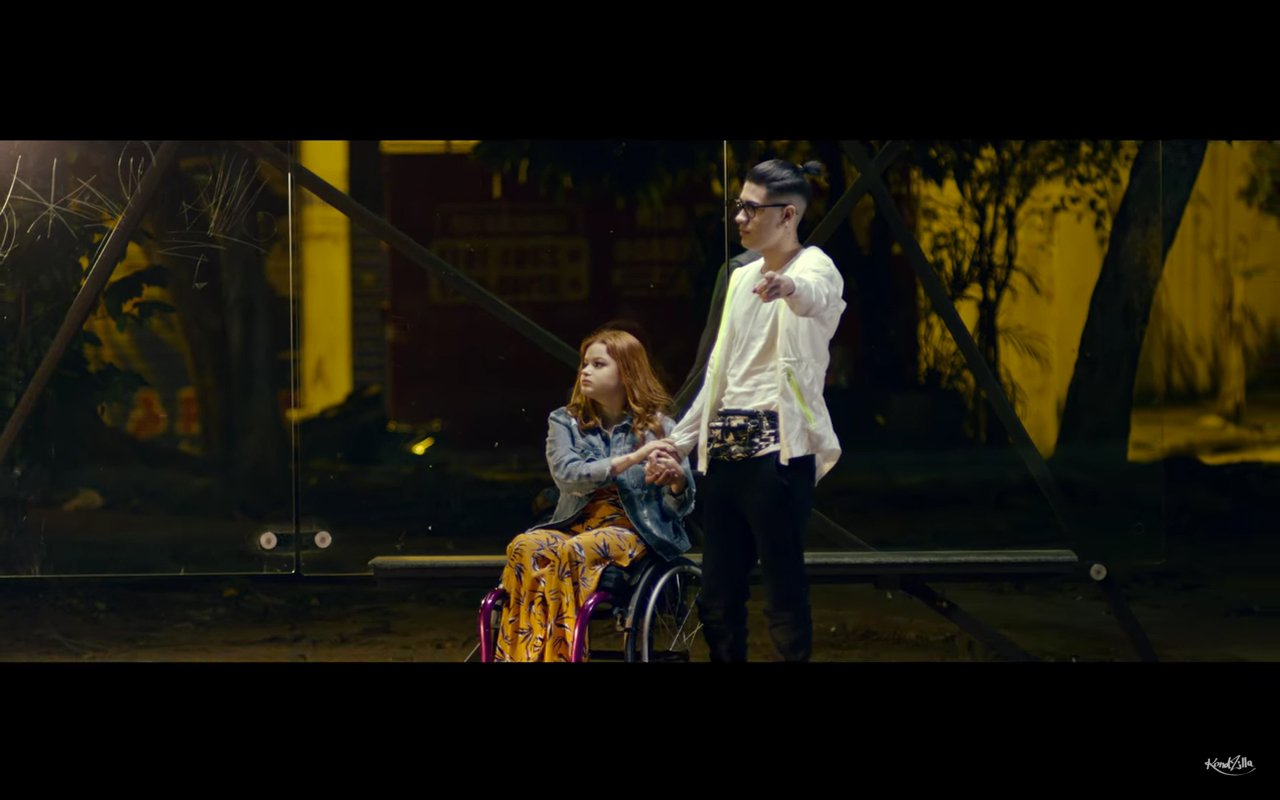 Descrição da imagem #PraCegoVer: Imagem no formato retangular, na horizontal. Cena do clipe de O Bebê, com Kevinho e MC Kekel. Kevinho está de mão dadas com a namorada, que é cadeirante, em um ponto de ônibus. Ele está dando sinal. Fim da descrição.