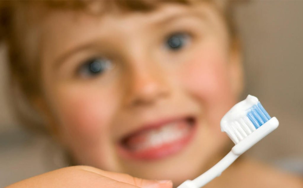Descrição da imagem #PraCegoVer: Imagem no formato retangular, na horizontal. Em primeiro plano, vemos uma escova de dentes com creme dental, e ao fundo, um pouco desfocado, uma menina que está sorrindo. Fim da descrição.