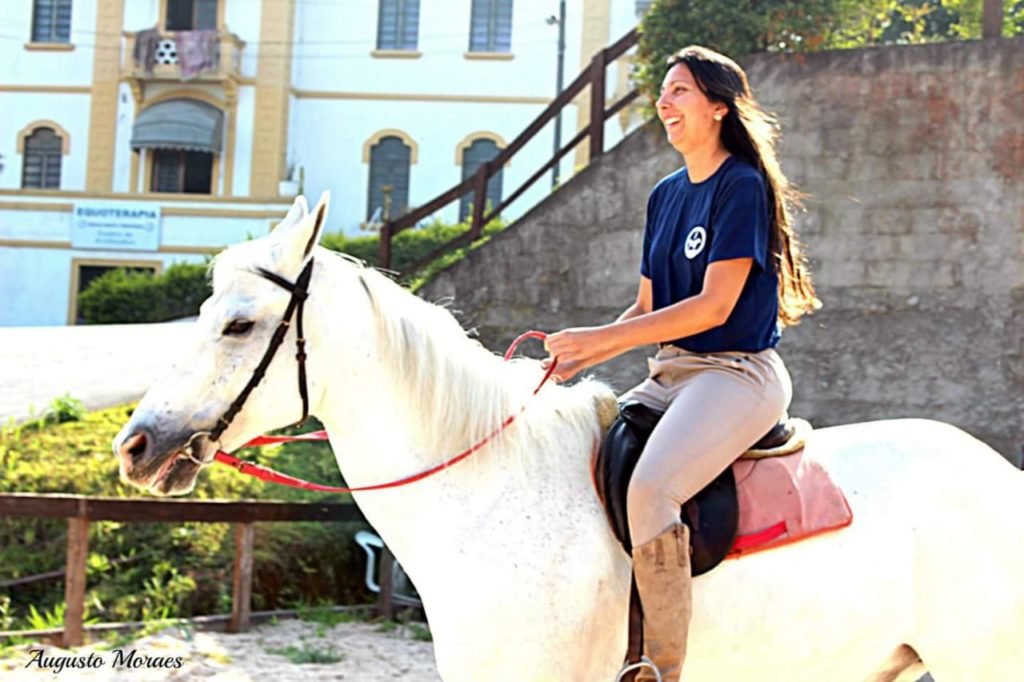 Descrição da imagem: foto da equoterapeuta Eliane montada no cavalo Raio, um cavalo branco. Eliane é uma mulher branca, que tem cabelos castanhos, lisos e compridos. Fim da descrição.