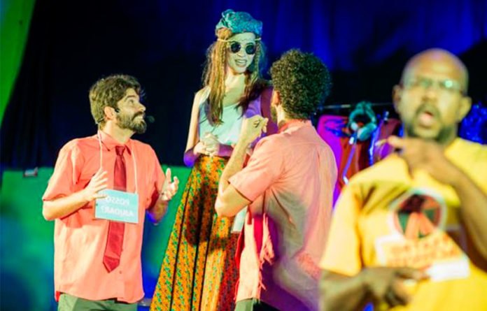 Descrição da imagem #pracegover: Foto de uma apresentação teatral com interpretação em Libras. Ao fundo vemos os atores no palco e em primeiro plano, vemos o intérprete de Libras, traduzindo a apresentação. Fim da descrição.