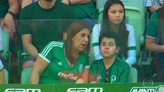 Descrição da imagem #PraCegoVer: Imagem no formato retangular, na horizontal. Imagem da torcida do Palmeiras. A mãe está sentada ao lado de seu filho, que é cego. Ela está descrevendo detalhes da partida para o menino. Eles estão usando a camisa verde, do Palmeiras. Fim da descrição.