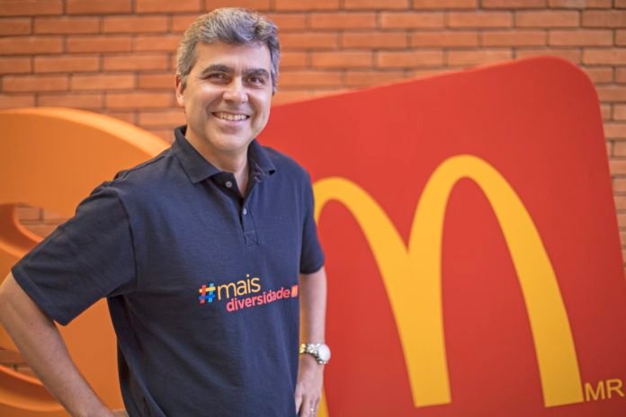 Descrição da imagem #PraCegoVer: Imagem no formato retangular, na horizontal. O diretor de RH do McDonald's, Marcelo Nóbrega. Marcelo é um homem moreno, de cabelos grisalhos. Ele usa uma camiseta azul marinho, onde está escrito #maisdiversidade, em letras coloridas. Marcelo está sorrindo, com as mãos na cintura, à frente do logo vermelho com um M amarelo, do McDonald's. Fim da descrição.