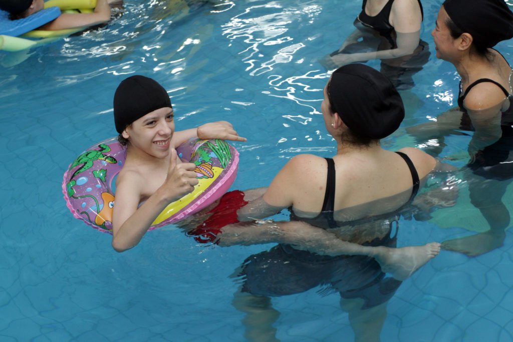 Descrição da imagem #PraCegoVer: Imagem no formato retangular, na horizontal. Um rapaz, com deficiência física, está em uma sessão de hidroterapia, dentro de uma piscina. Ele está acompanhado de uma mulher. Fim da descrição.