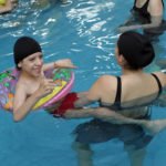 Descrição da imagem #PraCegoVer: Imagem no formato retangular, na horizontal. Um rapaz, com deficiência física, está em uma sessão de hidroterapia, dentro de uma piscina. Ele está acompanhado de uma mulher. Fim da descrição.