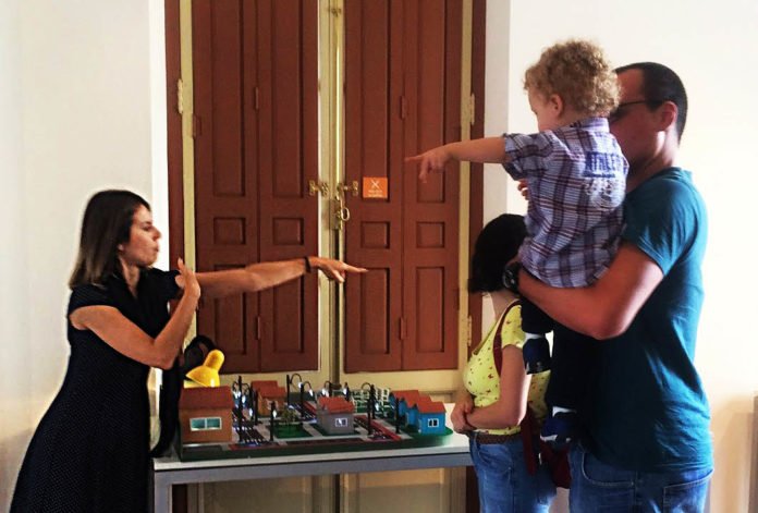 Descrição da imagem #PraCegoVer: Imagem no formato retangular, na horizontal. A educadora surda durante atendimento a visitantes no Museu da Energia. A mulher conversa, em Libras, com um homem que segura uma criança no colo. Fim da descrição.