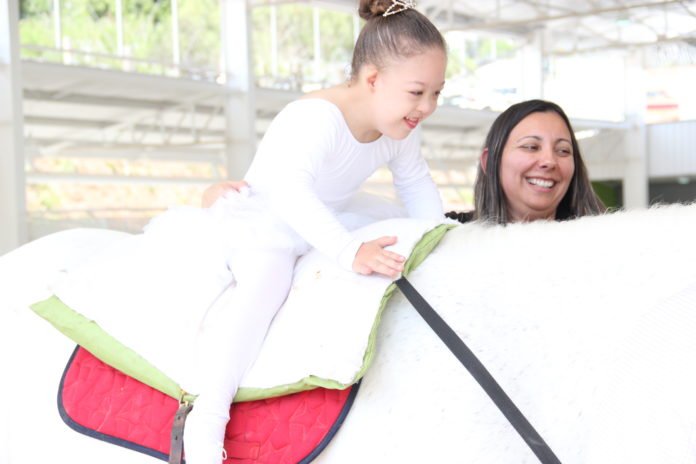 Descrição da imagem #PraCegoVer: Imagem no formato retangular, na horizontal. Uma menina, que tem síndrome de Down, está montada em um cavalo branco. Ao seu lado está a equoterapeuta Eliane Baatsch. A menina usa uma roupa branca. Elas estão na apresentação durante a inauguração do centro de equoterapia de Barueri. Fim da descrição.