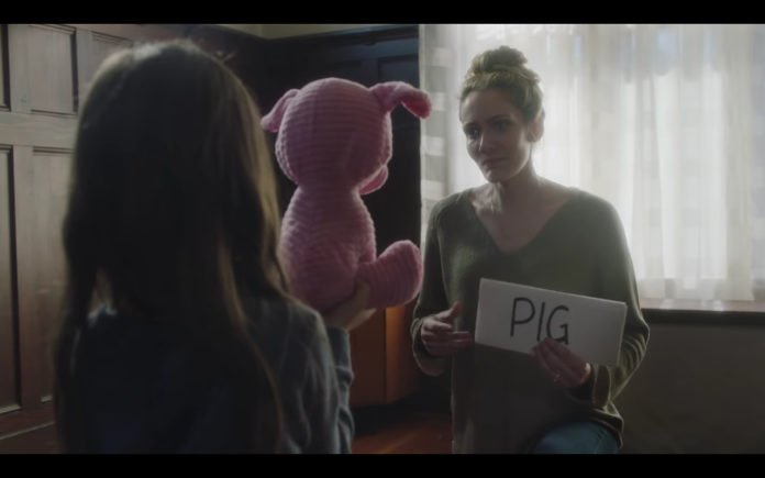 Descrição da imagem #PraCegoVer: Imagem no formato retangular, na horizontal. Cena do comercial. A menina está de frente para sua mãe. Ela segura seu porquinho rosa de pelúcia. A mãe está segurando um papel onde está escrito PIG, que significa porco. Fim da descrição.