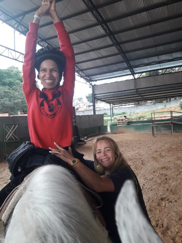 Descrição da imagem #PraCegoVer: Imagem no formato retangular, na vertical. Elaine, que sofreu um AVC, está montada em um cavalo branco. Ela está com os braços para cima, e sorri. Elaine usa uma blusa vermelha e uma calça preta. Ao seu lado está uma das profissionais da equipe de equoterapia. Fim da descrição.