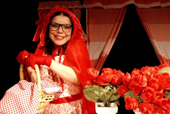 Descrição da imagem #PraCegoVer: Imagem no formato retangular, na horizontal. Cena da peça 'A Chapeuzinho Vermelho'. Nela vemos a protagonista, usando sua roupa vermelha e branca. Ela está segurando uma cesta. Ao fundo, vemos uma janela e a seu lado há vasos brancos com rosas vermelhas. Chapeuzinho Vermelho é uma menina morena, com cabelos pretos, lisos, e usa óculos. Ela está sorrindo. Fim da descrição.