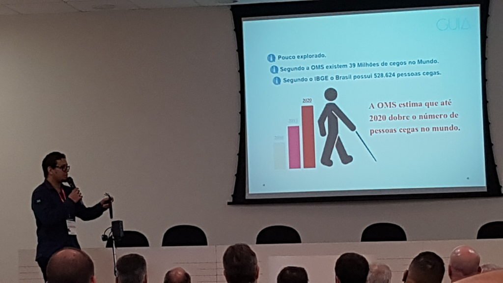 Descrição da imagem #PraCegoVer: Imagem no formato retangular, na horizontal. Apresentação do projeto de automação para bengalas. Um telão com a imagem de um ícone com uma bengala e alguns dados. Pessoas acompanham a apresentação, que está sendo feita por um homem. Fim da descrição.