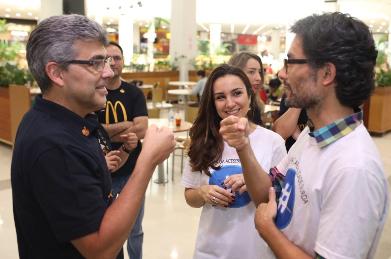 Descrição da imagem #PraCegoVer: Imagem no formato retangular, na horizontal. O diretor de RH do McDonald's, Marcelo Nóbrega, está aprendendo alguns sinais em Libras com o arquiteto Alexandre Ohkawa,. Ao fundo, a jornalista Milena Machado Grupo acompanha tudo. Fim da descrição.