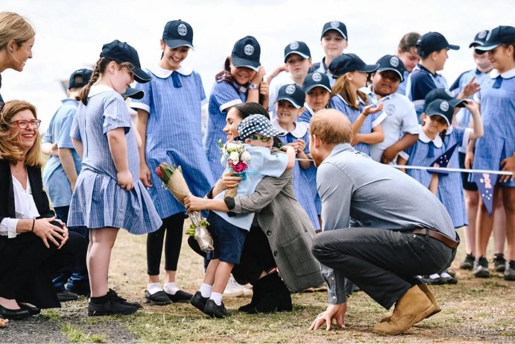 Descrição da imagem #PraCegoVer: Imagem no formato retangular, na horizontal. Meghan sendo abraçada pelo menino Luke, que tem síndrome de Down. Ao seu lado está Harry e a comitiva do casal real, durante visita à Dubbo, na Austrália. Fim da descrição.
