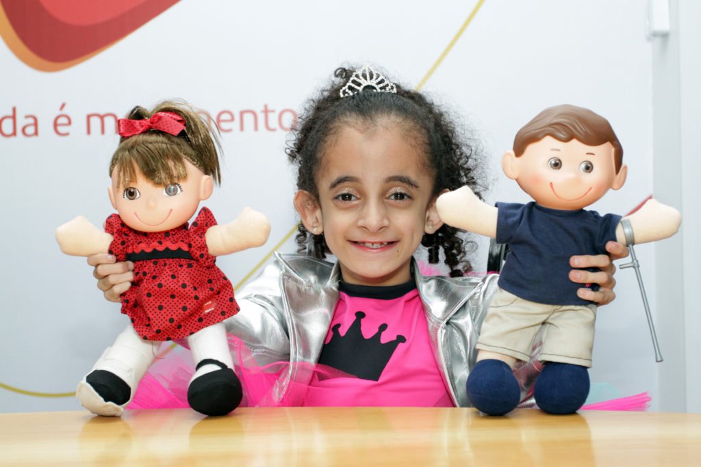 Descrição da imagem #PraCegoVer: Imagem no formato retangular, na horizontal. A menina Luara Crystal, criança símbolo do Teleton 2018. Ela.é uma menina morena e tem cabelos cacheados. Luara está segurando os mascotes do Teleton, Tonzinho e Nina. Luara sorri. Fim da descrição.