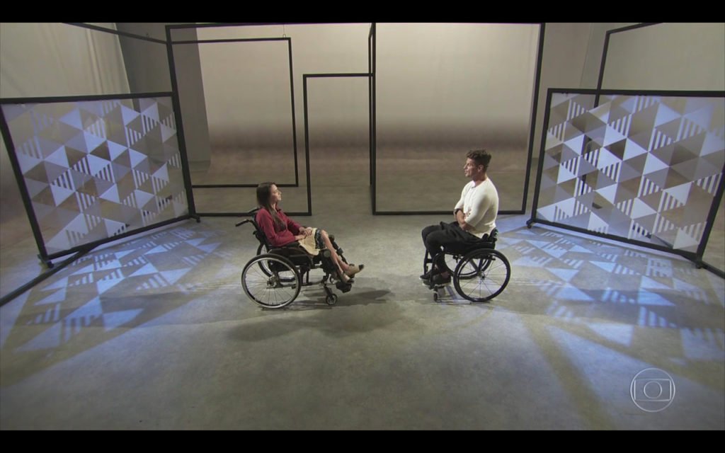 Descrição da imagem #PraCegoVer: Imagem no formato retangular, na horizontal. Cena do quadro 'Você por aqui', do Fantástico. Laís Souza e Fernando Fernandes estão frente a frente. Ambos são cadeirantes. Laís usa uma blusa cor vinho e tem um chale bege sobre as pernas. Fernando usa uma calça preta e uma camiseta branca. Fim da descrição.