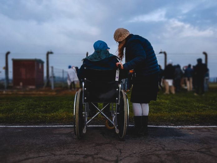 Descrição da imagem #PraCegoVer: Imagem no formato retangular na horizontal. Duas pessoas estão de costas para a câmara e conversam. Uma delas é cadeirante e a outra está de pé. Fim da descrição.