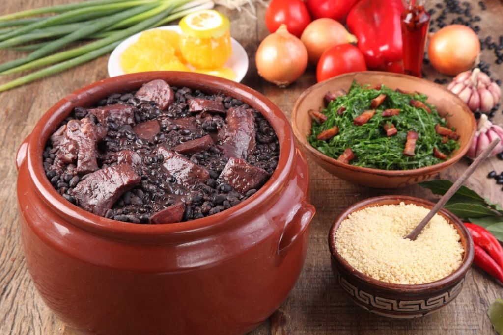 Descrição da imagem #PraCegoVer: Uma mesa com alguns dos pratos que compõem uma feijoada. São eles feijão preto com carne seca e paio, couve, farofa e laranja. Fim da descrição.