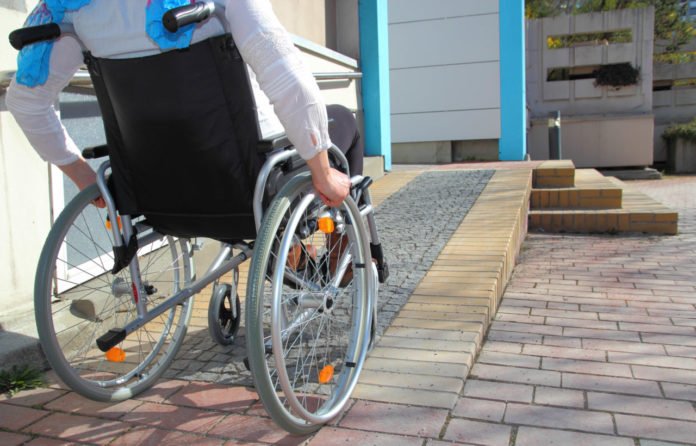 Descrição da imagem #PraCegoVer: Imagem no formato retangular, na horizontal. Uma rampa em uma calçada. Uma pessoa está sentada em uma cadeira de rodas, subindo a rampa. Fim da descrição.