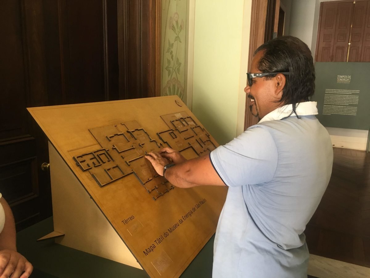 Descrição da imagem #PraCegoVer: Imagem no formato retangular, na horizontal. O jornalista Ismael da Silva, que é cego, está tateando o mapa tátil do Museu da Energia de São Paulo. Ismael é um homem moreno e tem cabelos pretos na altura dos ombros. Ele está usando óculos escuros e uma camisa polo azul com gola branca. Ele está sorrindo. Fim da descrição.