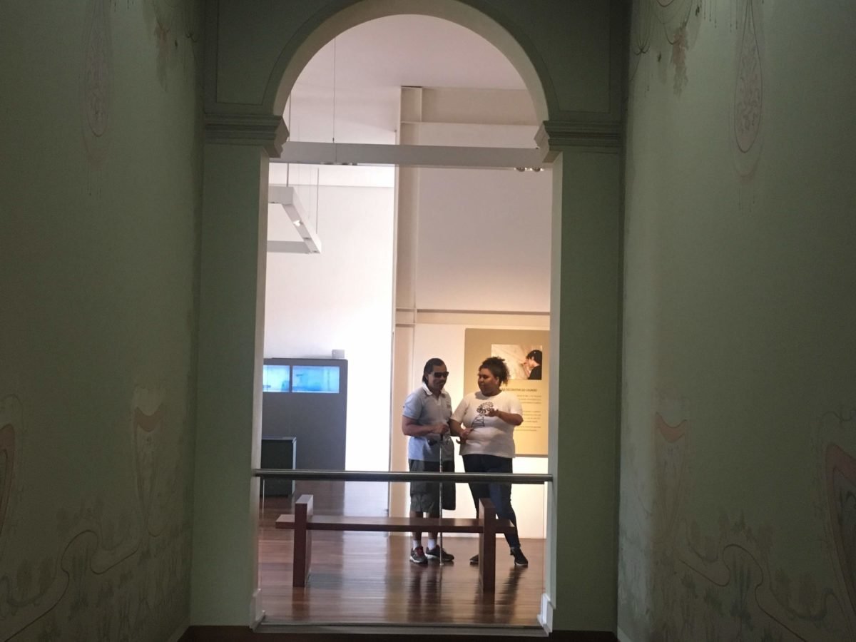 Descrição da imagem #PraCegoVer: Imagem no formato retangular, na horizontal. O jornalista Ismael da Silva, que é cego, está em uma ala do Museu da Energia de São Paulo, ao lado da educadora do museu. Eles estão envoltos a uma moldura formada pelas paredes do museu. Fim da descrição.