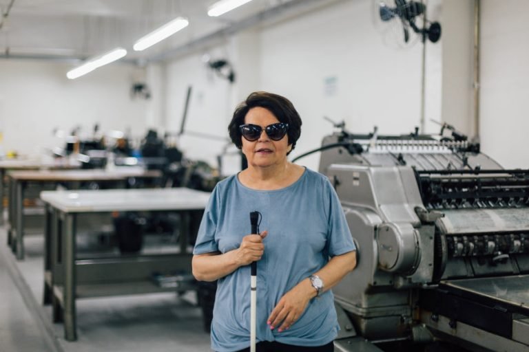 Descrição da imagem #PraCegoVer: Imagem no formato retangular na horizontal. A coordenadora da Fundação Dorina, Regina Oliveira. Ela está na gráfica braile da fundação. Regina é uma mulher morena, de pele clara e cabelos pretos, lisos, na altura dos ombros. Ela está segurando sua guia. Fim da descrição.