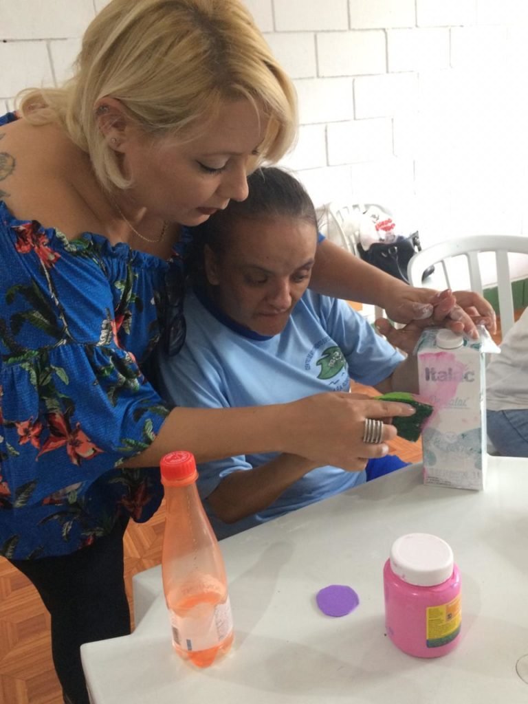 Descrição da imagem #PraCegoVer: Psicopedagoga durante atendimento a um paciente com deficiência intelectual. Juntas, elas estão fazendo uma arte. Fim da descrição.