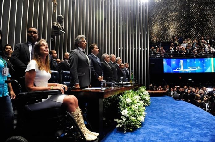 Descrição da imagem #PraCegoVer: Imagem no formato retangular, na horizontal. A senadora Mara Gabrilli está na plenária do Senado, durante sessão solene. Mara está ao lado de outros senadores e convidados da solenidade. Mara usa um vestido branco e uma bota bege. Ela está com os cabelos soltos. Mara é uma mulher morena, de pele clara e tem cabelos lisos e compridos. Fim da descrição.