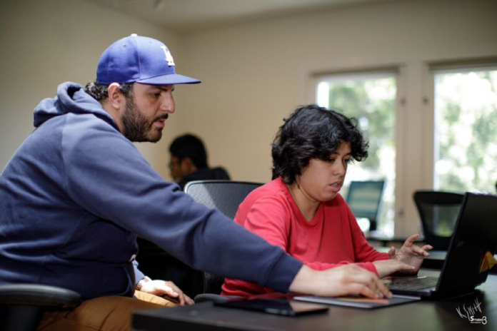 Descrição da imagem #PraCegoVer: Imagem no formato retangular, na horizontal. Um jovem está sentado na frente do computador e recebe orientações de um homem que está ao seu lado. Fim da descrição.