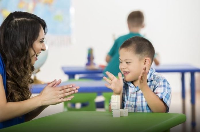 #descriçãodaimagem: Foto de um menino com síndrome de Down. Ele está sentado em sua carteira, na escola, ao lado de uma educadora. Eles estão batendo palmas. Ao fundo, estão outras crianças. Fim da descrição.