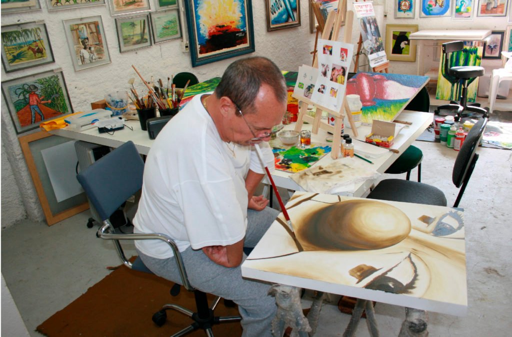 Descrição da imagem #PraCegoVer: A imagem está no formato retangular, na horizontal. Um homem com deficiência física está segurando um pincel com a boca e pinta um quadro. Fim da descrição.
