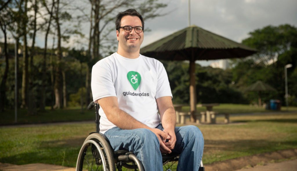 Descrição da imagem #PraCegoVer: Bruno Mahfuz, criador do aplicativo Guiaderodas. Bruno é um homem moreno, de pele clara e cabelos pretos, curtos. Bruno está sentado em sua cadeira de rodas e usa uma camiseta com o logo do app Guiaderodas. Fim da descrição.