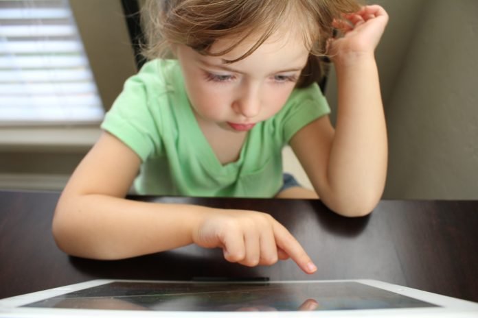 Descrição da imagem #pracegover: Uma menina está sentada em uma mesa, usando um tablet. Ela tem aproximadamente cinco anos e está vestindo uma blusa verde. Fim da descrição.