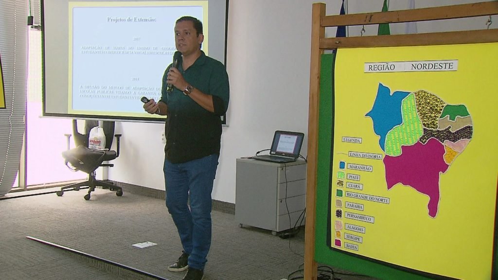 Descrição da imagem #PraCegoVer: O professor que criou os mapas táteis durante apresentação de seu projeto. Ele está em uma sala de aula e segura um microfone. Ao seu lado está um dos mapas com textura criados por ele para ensinar alunos com deficiência visual. Fim da descrição.