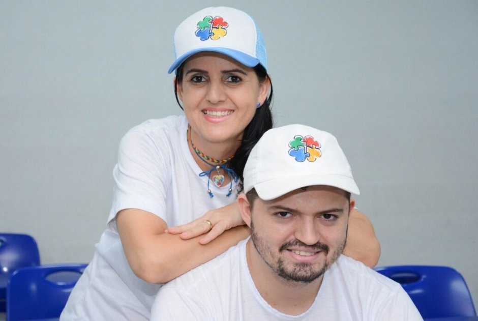 Descrição da imagem #PraCegoVer: Imagem no formato retangular, na horizontal. O jovem médico Enã Rezende com sua mãe Érica. Ele está sentado e ela está atrás dele, com os braços apoiados em suas costas. Os dois estão com camiseta branca e boné branco com um desenho colorido. Fim da descrição.