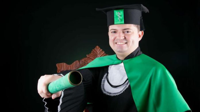 Descrição da imagem #PraCegoVer: Imagem no formato retangular, na horizontal. O jovem médico Enã Rezende durante sua formatura. Ele está usando uma beca preta com capa verde e segura o canudo do diploma com a mão direita. Enã é um rapaz moreno, dEle está sorrindo. Fim da descrição.