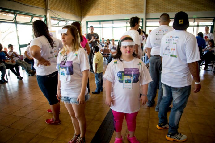 Descrição da imagem #PraCegoVer: Imagem no formato retangular, na horizontal. Um grupo de jovens durante o treinamento da Escola de Gente. Vemos, em destaque, uma moça com síndrome de Down. Todos estão com um papel colado na testa. Fim da descrição.
