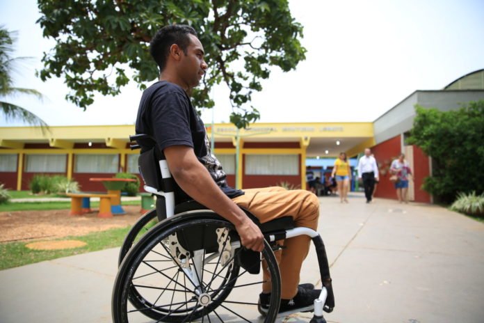 Descrição da imagem #PraCegoVer: Imagem no formato retangular, na horizontal. Um rapaz com deficiência física está em sua cadeira de rodas. A imagem mostra o rapaz de lado. Ele é moreno e tem cabelos pretos, curtos. Ele está usando uma camiseta preta e uma calça cor mostarda. Fim da descrição.