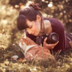 Descrição da imagem #pracegover: Foto da fotógrafa Sabine com o gato Kazuo. Eles estão em meio a um jardim, com muitas flores. Kazuo está deitado, na frente de Sabine, que também está deitada no chão. Kazuo é um gato branco e laranja e Sabine é uma mulher morena, de pele clara, e cabelos lisos, pretos, que estão amarrados. Ela segura sua câmera enquanto olha para o gato. Fim da descrição.