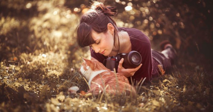 Descrição da imagem #pracegover: Foto da fotógrafa Sabine com o gato Kazuo. Eles estão em meio a um jardim, com muitas flores. Kazuo está deitado, na frente de Sabine, que também está deitada no chão. Kazuo é um gato branco e laranja e Sabine é uma mulher morena, de pele clara, e cabelos lisos, pretos, que estão amarrados. Ela segura sua câmera enquanto olha para o gato. Fim da descrição.