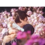 Descrição da imagem #PraCegoVer: Imagem no formato retangular, na horizontal. A fotógrafa Sabine com o gato Kazuo. Eles estão em meio a um jardim, com muitas flores. Kazuo está no colo de Sabine, que está beijando o gato. Fim da descrição.