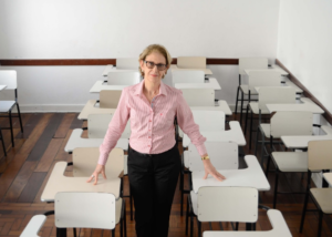 Descrição da imagem #PraCegoVer: Imagem no formato retangular, na horizontal. A diretora Lenice Micheletti está em uma sala de aula vazia. Ela está entre as carteiras. Lenice é uma mulher de pele clara e cabelos loiros, curtos. Ela está usando uma camisa de mangas compridas cor de rosa e uma calça social preta. Ela está sorrindo. Fim da descrição.
